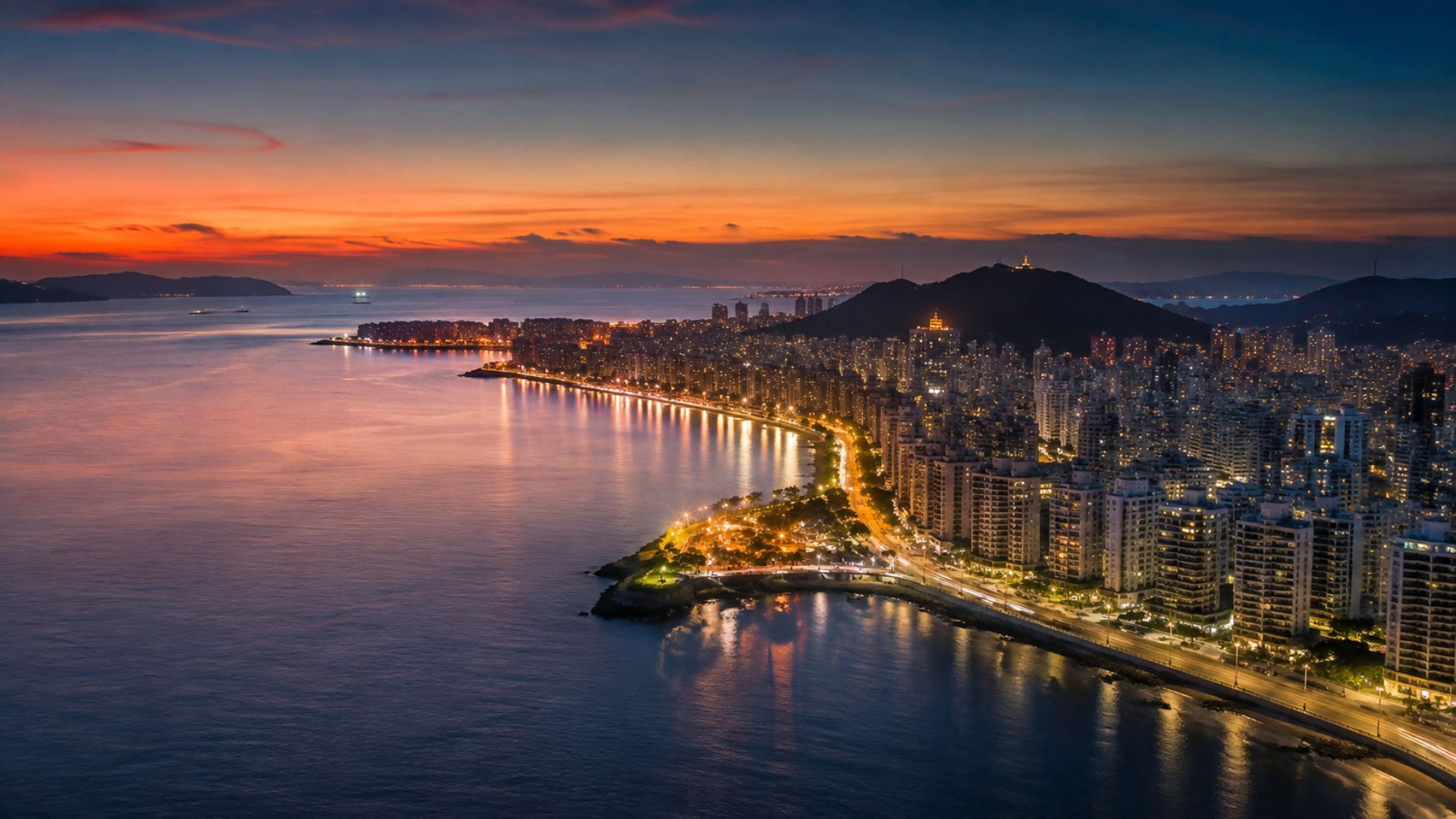 Vista aérea panorâmica de uma orla urbana ao entardecer, com prédios altos iluminados à beira-mar, luzes da avenida refletindo na água, céu em tons de laranja e azul profundo, montanhas ao fundo, fotografia realista aérea de drone