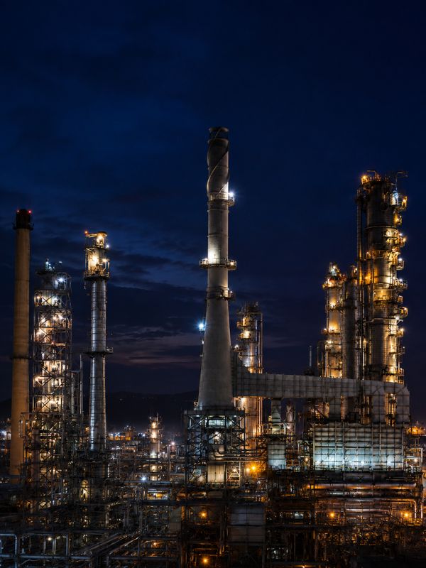 Vista noturna de uma refinaria industrial com torres metálicas e chaminés iluminadas em tons dourados sobre céu azul profundo, estrutura de tubulações e escadarias, fotografia realista industrial